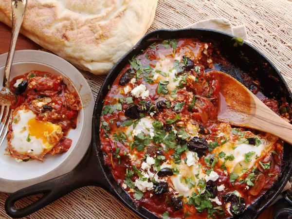 Shakshuka in a cast iron skillet, topped with parsley and olives, with a scooped single serving in a bowl and pita bread on the side.